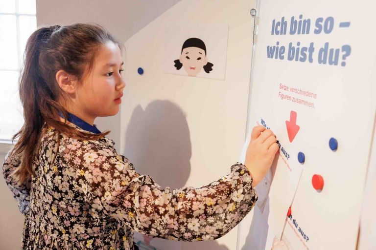 Ein M&auml;dchen schreibt an eine Wand auf der steht 'Ich bin so. Wie bist Du?'